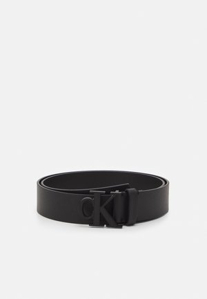 Ceinture en cuir noir roulée avec une boucle à logo "cK" noire mate centrée sur un fond gris clair uni.