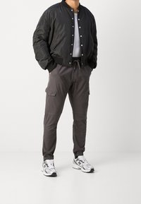 Veste bomber noire, t-shirt à col rond blanc, pantalon cargo gris avec poches, et chaussures de sport blanches avec des accents noirs. Design simple et fonctionnel.