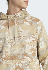 Sudadera beige con patrón de camuflaje, hecha de una tela suave, con un gran logo de Adidas en relieve y capucha con cordón.