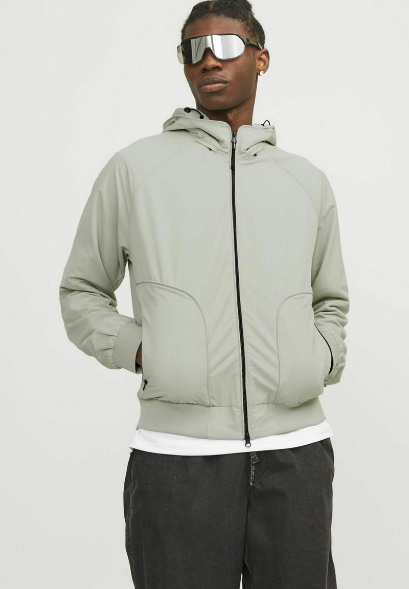Light gray zip-up jacket with a hood, elastic cuffs, and side pockets. Worn over a white shirt, paired with mirror sunglasses.