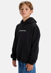 Černá mikina s volným střihem, se přední kapsou a bílým logem na hrudi. Měkká tkanina se hladkým povrchem a kapucí na šňůrku.