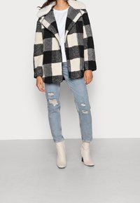 Frau trägt eine schwarz-weiße karierte Fleecejacke, ein weißes Hemd, hellblaue, zerrissene Jeans und weiße, hochhackige Knöchelstiefel vor einem einfarbigen Hintergrund.