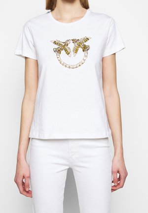 Biała koszulka z krótkim rękawem z dwoma złotymi i bursztynowymi pszczołami z cekinów na kropkowanym, okrągłym wzorze na klatce piersiowej, noszona z białymi spodniami.