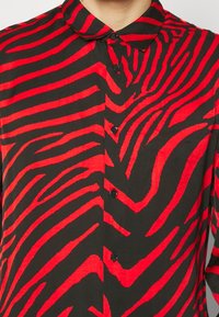 Camicia a bottoni rossa e nera con motivo a strisce zebrate. Tessuto liscio con finitura lucida, colletto al collo e bottoni neri.