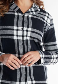 Vrouw in een zwart, wit en grijs geruit overhemd met lange mouwen, met de handen zachtjes gekruist op taillehoogte.