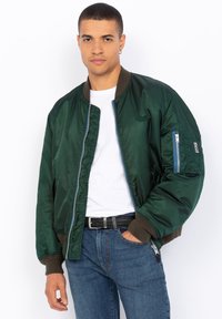 Giacca bomber in raso verde con polsini e vita elasticizzati, dotata di una tasca con zip sulla manica, indossata sopra una maglietta bianca e un paio di jeans.