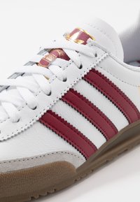 Vita lädersneakers med tre vinröda ränder, en grå mocka tådel och en texturerad brun gummisula. Guldaccent på plösen.