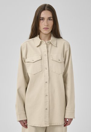 Žena s dlhými hnedými vlasmi, oblečená v béžovej košeli s gombíkmi a dvoma náprsnými vreckami na jednoduchom sivom pozadí.