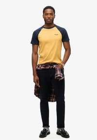 Camiseta amarilla de manga corta con mangas raglán en azul marino, pantalones vaqueros negros y una camisa de cuadros atada a la cintura. Zapatos negros con suela plana.