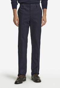 Navyblaue Hosen aus glattem Stoff, mit geradem Bein und Bundfalten. Getragen mit dunkelbraunen Lederschuhen.