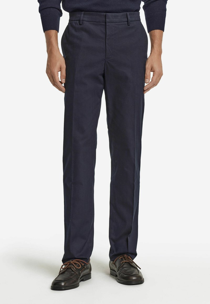 Navyblaue Hosen aus glattem Stoff, mit geradem Bein und Bundfalten. Getragen mit dunkelbraunen Lederschuhen.