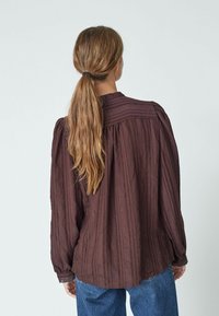 Tumepruun pluus vertikaalsete triipudega, pikkade varrukatega, volditud detailidega õlgadel ja pitsaktsentidega mansettides. Vabakujundus ümmarguse kaelusega.