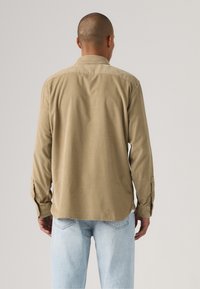 Tan longsleeve shirt met een button-down kraag, met een zachte textuur en een ronde zoom. Gekleed met lichtblauwe jeans, gezien van de achterkant.