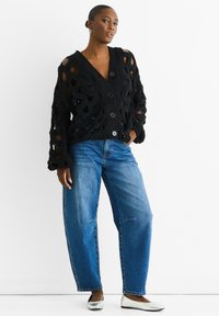 Black knit cardigan with large open weave and six buttons, paired with light blue relaxed-fit jeans and silver flats.