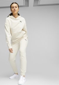 Sudadera crema con bolsillo canguro y logo negro; combinada con pantalones jogger a juego. Zapatillas blancas completan el look. Textura suave en toda la prenda.