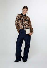 Blusa in satin con stampa leopardata e dettagli neri, abbinata a jeans scuri a gamba larga. Il modello tiene una borsa a tracolla bianca.