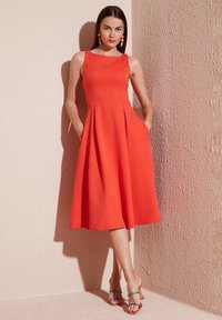 Röd ärmlös klänning med figurnära liv, utsvängd kjol och sidofickor. Texturerad tyg kompletterar den minimalistiska designen.