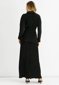 Black pleated dress with long sleeves, a collar, and a flowing hem. The fabric features vertical ribbing and a belted waist.