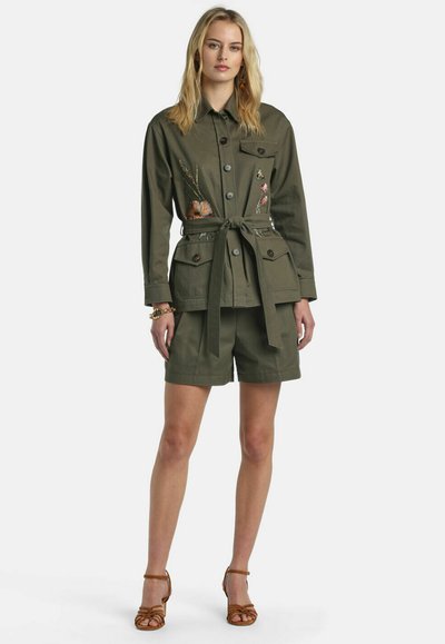 Woman wearing olive green belted jacket with floral embroidery, matching shorts, and tan strappy heels, standing against white background.