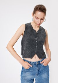 Grijze kortlopende vest met een ronde hals, knopen aan de voorkant en een licht gestructureerde afwerking. Gecombineerd met blauwe high-waisted jeans.