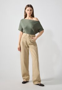 Top oversized en maille côtelée verte avec encolure off-shoulder associé à un pantalon large taille haute beige et des ballerines noires.