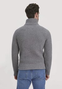 Grauer Strickpullover mit hohem Kragen und geripptem Saum, der ein strukturiertes Muster und angepasste lange Ärmel hat. Getragen über blauen Jeans.