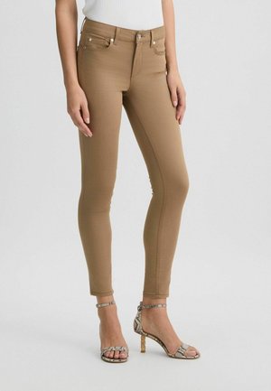 Pantalon slim couleur camel porté avec un top blanc et des sandales à talons hauts ouvertes en peau de serpent sur un fond clair uni.