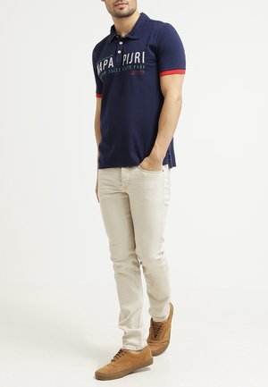 Polo marine avec des accents rouges, comportant un logo et une impression de texte, associé à un jean beige clair et des baskets marron.