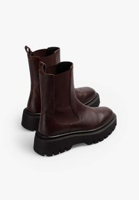 Botas Chelsea de cuero marrón con laterales elásticos, punteras redondeadas y una suela de goma negra gruesa y texturizada. Cuenta con trabillas para facilitar el uso.