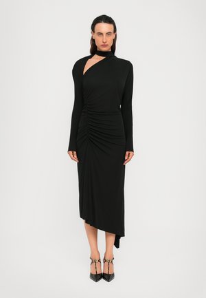 Woman stands in a black asymmetrical long-sleeve dress with ruched detailing and cutout shoulder, wearing black pointed-toe heels with straps.