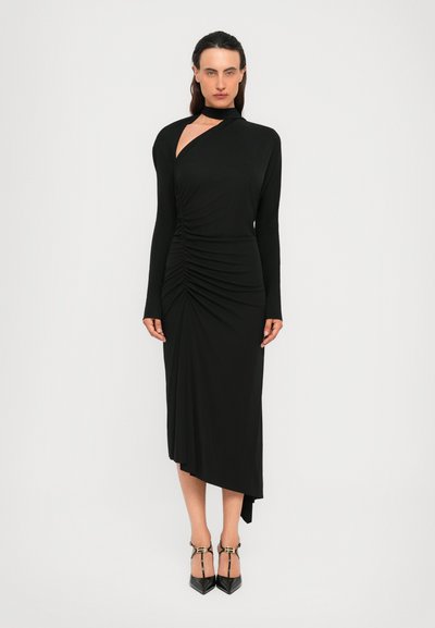 Une femme se tient dans une robe noire asymétrique à manches longues avec des détails froncés et une épaule dénudée, portant des escarpins noirs à bout pointu avec des bretelles.