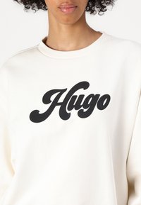 Vaalea kermaisen värinen collegepaita, jossa on pyöreä kaula-aukko ja musta "Hugo"-logo tyylitellyllä fontilla rinnan ylle. Pehmeää kangasta ja rento istuvuus.