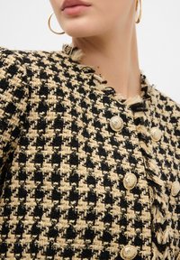 Svart och beige houndstooth tweedjacka med guldknappar, fransade kanter och en strukturerad design. Nära vy framhäver textur och detaljer.