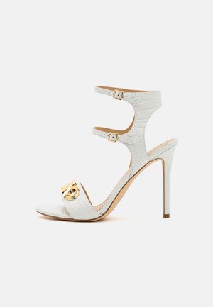 Sandale blanche à talon haut avec texture crocodile, deux brides chevilles avec boucles dorées et une boucle décorative dorée sur la bride des orteils.