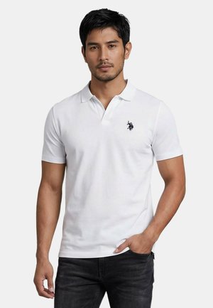 Mann trägt weißes kurzärmeliges Poloshirt mit schwarzem besticktem Logo, schwarze Jeans, steht mit einer Hand in der Tasche vor einfarbigem Hintergrund.