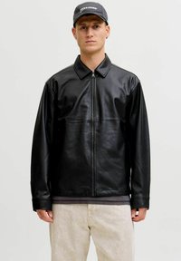 Schwarze Lederjacke mit Reißverschluss vorne, spitzem Kragen und langen Ärmeln. Getragen über einem grauen Hemd, kombiniert mit hellfarbigen Hosen.