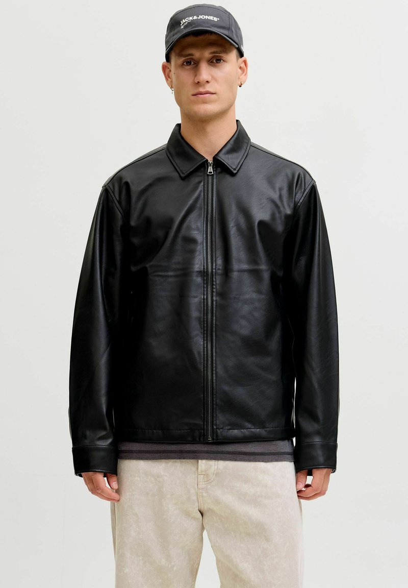 Schwarze Lederjacke mit Reißverschluss vorne, spitzem Kragen und langen Ärmeln. Getragen über einem grauen Hemd, kombiniert mit hellfarbigen Hosen.