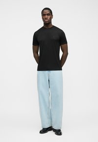 Noormees seisab otse vaataja poole, kandes musta lühikeste varrukatega t-särki, helerohelisi laia säärega teksapükse ja musti jalanõusid, taust on lihtne.