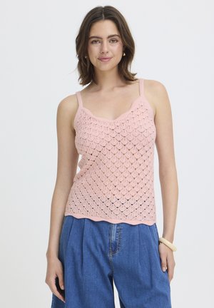 Jeune femme portant un haut sans manches rose en crochet et un pantalon ample en jean bleu, posant devant un fond neutre.