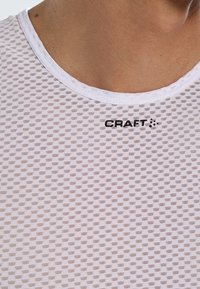Maglietta bianca traspirante con un motivo a rete testurizzata. Presenta un collo rotondo e un logo nero "CRAFT" sul petto.