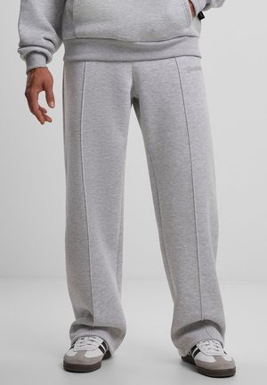 Mann trägt hellgraue Jogginghose und passendes Sweatshirt mit weißen und grauen Turnschuhen, steht vor einem einfarbigen Hintergrund.