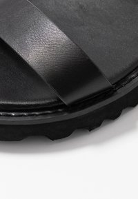 Musta sandaali, jossa on kaksi leveää hihnaa tekstuurimateriaalista, paksu, uritettu pohja ja näkyvät ompeleet reunan ympärillä.