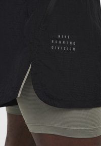 Čierna ľahká bežeck�á bunda s textúrovaným povrchom, so bielym logom "Nike Running Division", v kombinácii s olivovozelenými kompresnými šortkami.