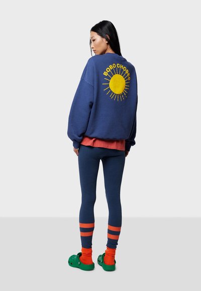 Mörkblå sweatshirt med gul solgrafik på ryggen, lager-på-lager över röd tröja, kombinerad med blå leggings och gröna träskor med orange strumpor.