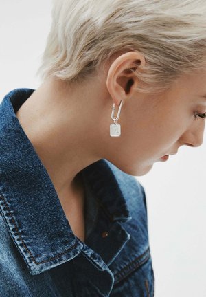Persona con cabello rubio corto que lleva un pendiente rectangular de plata y una chaqueta de denim azul, mirando ligeramente hacia abajo y a la derecha.
