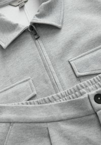 Giacca zip grigia chiara con colletto e due tasche sul petto, abbinata a pantaloni grigi coordinati con vita elastica e bottone nero.