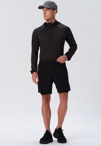 Lätt svart zip-up hoodie med raglanärmar, i kombination med svarta shorts. Modell bär en grå keps och svarta sneakers. Slät textur.