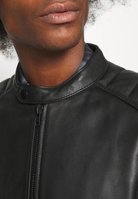 Gros plan d'une personne portant une veste en cuir noire avec une fermeture éclair et un col à bouton-pression, montrant les lèvres et une partie du cou.