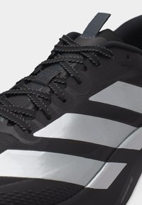 Κοντινή λήψη μαύρου αθλητικού παπουτσιού με μαύρα κορδόνια και ασημί διαγώνιες ρίγες στο πλάι, που δείχνει υφασμένο πέλμα και θηλιά έλξης.