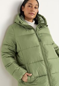 Kvinde iført en lys grøn dunjakke med hætte og en hvid turtle neck, står med den ene hånd i lommen mod en hvid væg.